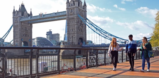 Three individuals walk along a riverside path with Tower Bridge in the background. The sky is clear and sunny, and trees are visible to the right.