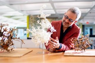A person in a red jacket smiles and examines a white intricate sculpture on a table. Two other sculptures, one made of wood and another resembling a cluster of geometric shapes, are displayed nearby in a well-lit room.