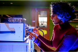 Person in a red sweater operates scientific equipment in a lab. The room is illuminated by blue lighting, and the background shows a door with a window. The person is focused on adjusting the machine's components.