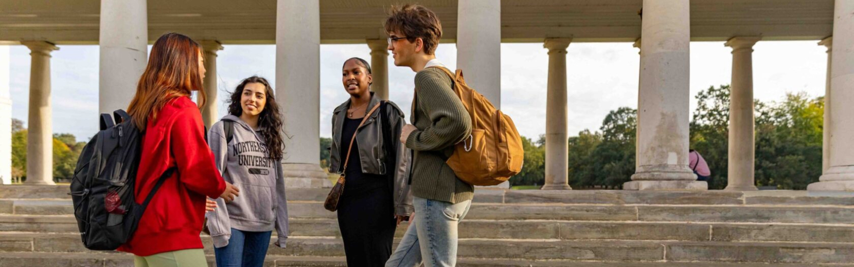 Four students stand in conversation near a set of wide stone steps and large columns. Two wear backpacks, and all are dressed casually. Trees are visible in the background under a clear sky.