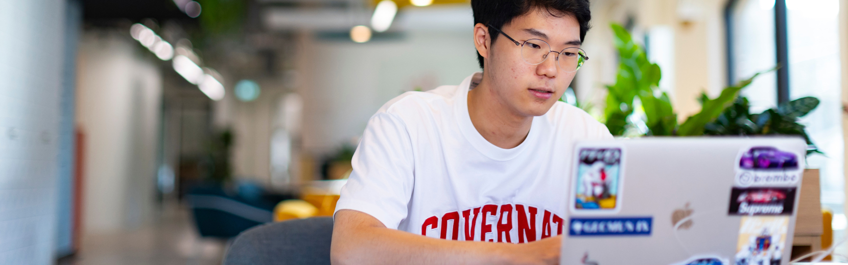 A person wearing glasses and a white T-shirt with red text is sitting at a table using a laptop covered in stickers. The background shows an open, modern office space with plants and seating areas.