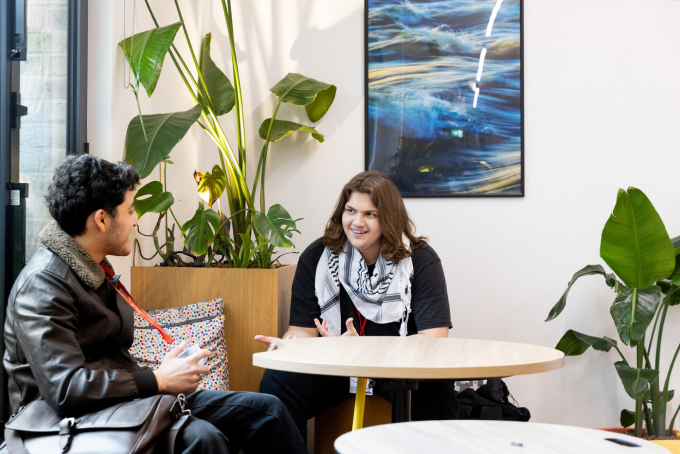 Two people sit at a round table in a bright room with plants and modern art on the wall. One is wearing a leather jacket and the other a scarf. They're engaged in conversation, creating a relaxed atmosphere.