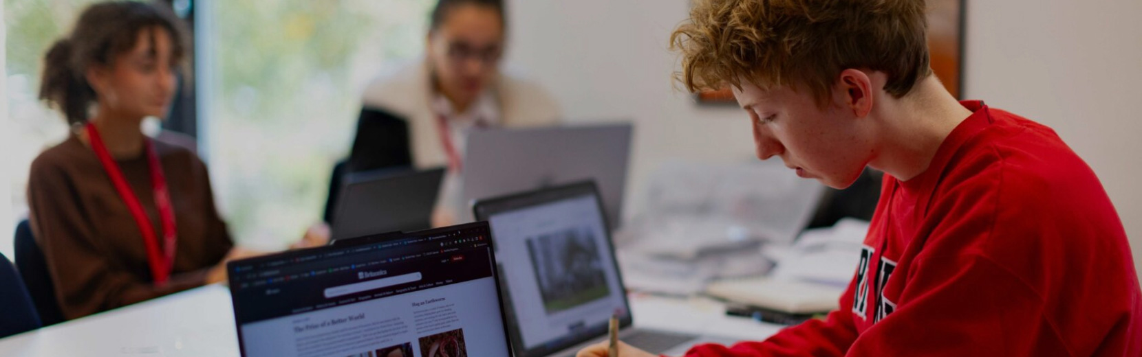 A young man in a red sweater is focused on writing at a desk with a laptop. Two other people are sitting in the background, working on their laptops. The room has a bright atmosphere with natural light coming from a window.