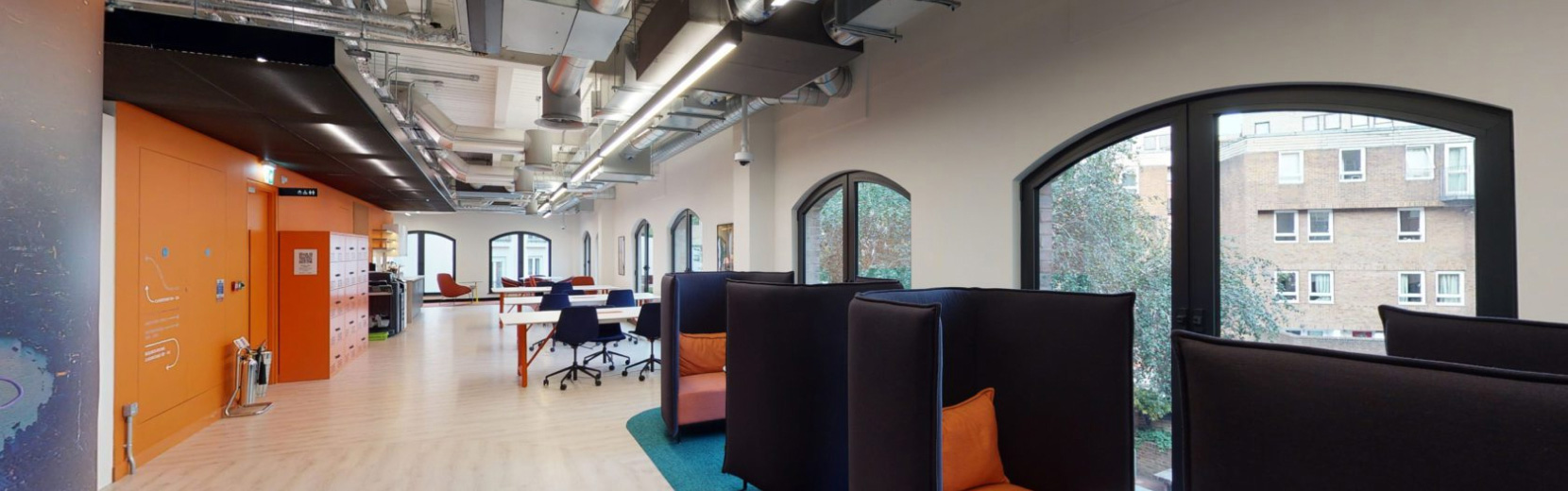 A modern office space with high ceilings, exposed ductwork, and large arched windows. The room is equipped with black high-backed chairs, desks, and orange accents. A row of windows overlooks a building and trees outside.