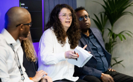 A woman with long curly hair speaks while holding papers, seated between two men. The man on the right wears glasses and a blue jacket. They are part of a panel discussion, with a screen and a plant in the background.