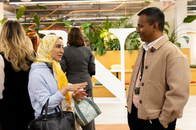 Two people are engaged in conversation at a social event indoors. One person is wearing a headscarf and holding a bag, while the other wears a textured jacket. They are surrounded by other attendees and greenery in the background.