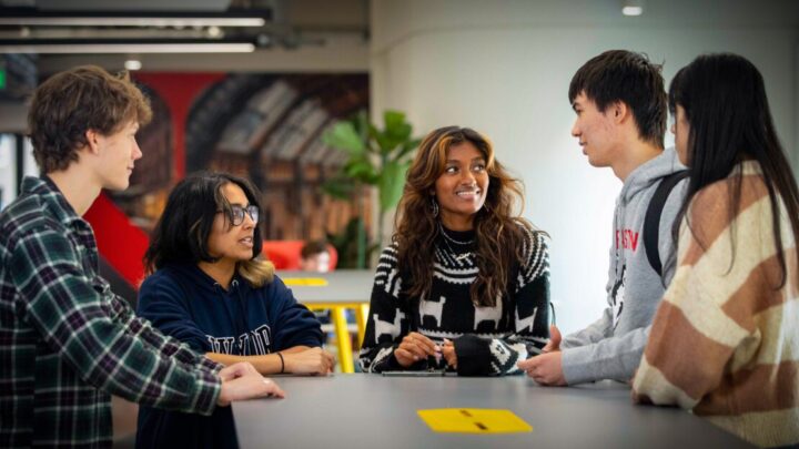 A diverse group of five young adults engaged in conversation around a table indoors. They are smiling and appear to be in a casual, social setting. The background shows a modern interior with colorful decor.