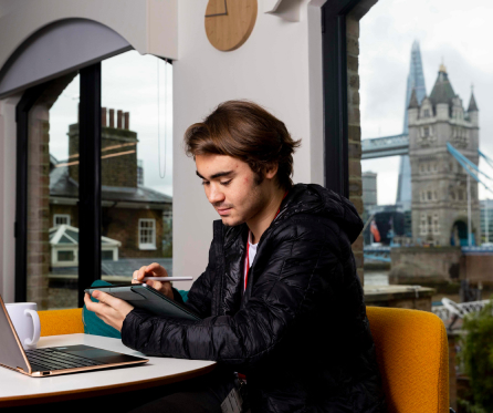 A person in a black jacket sits at a table with a digital tablet and stylus, working next to an open laptop. In the background, a large window reveals a view of Tower Bridge and the Shard in London.