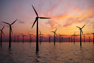 A wind farm with numerous wind turbines stands in the ocean under a vibrant sunset sky, with hues of orange, pink, and purple reflecting on the water's surface.