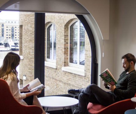 Two people are sitting in red chairs by a large arched window, reading books. The window overlooks brick buildings and water. The interior has neutral tones, creating a cozy atmosphere.