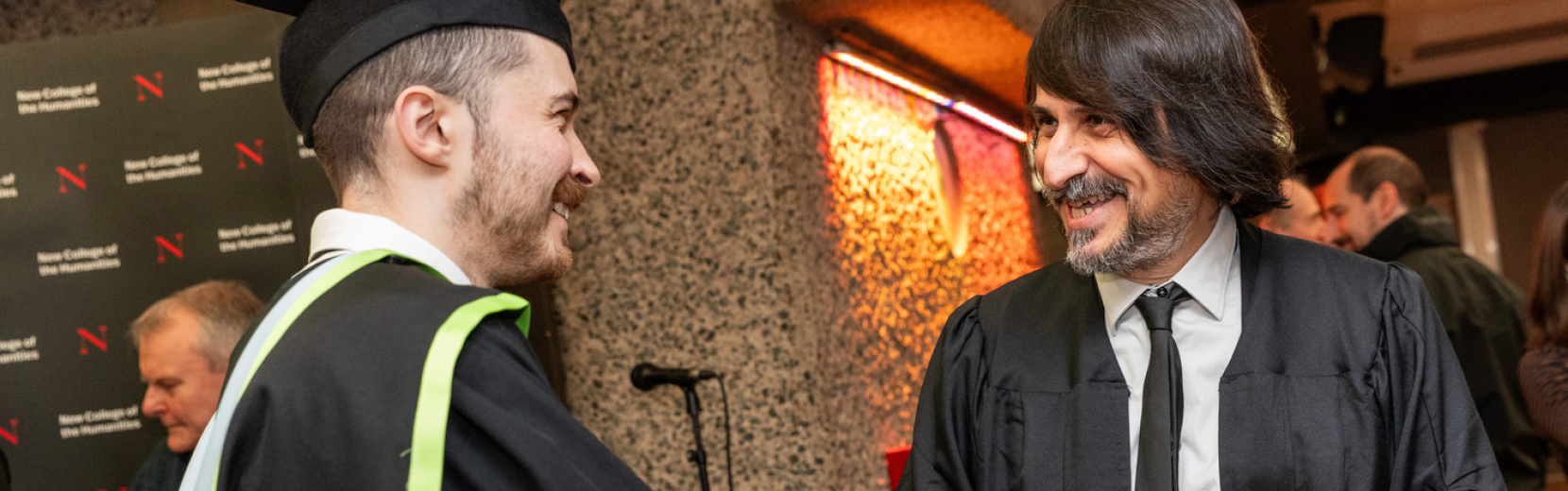 A graduate in a cap and gown shakes hands with a bearded man in a suit. They are smiling, standing in front of a textured wall with a logo in the background, during a formal event.