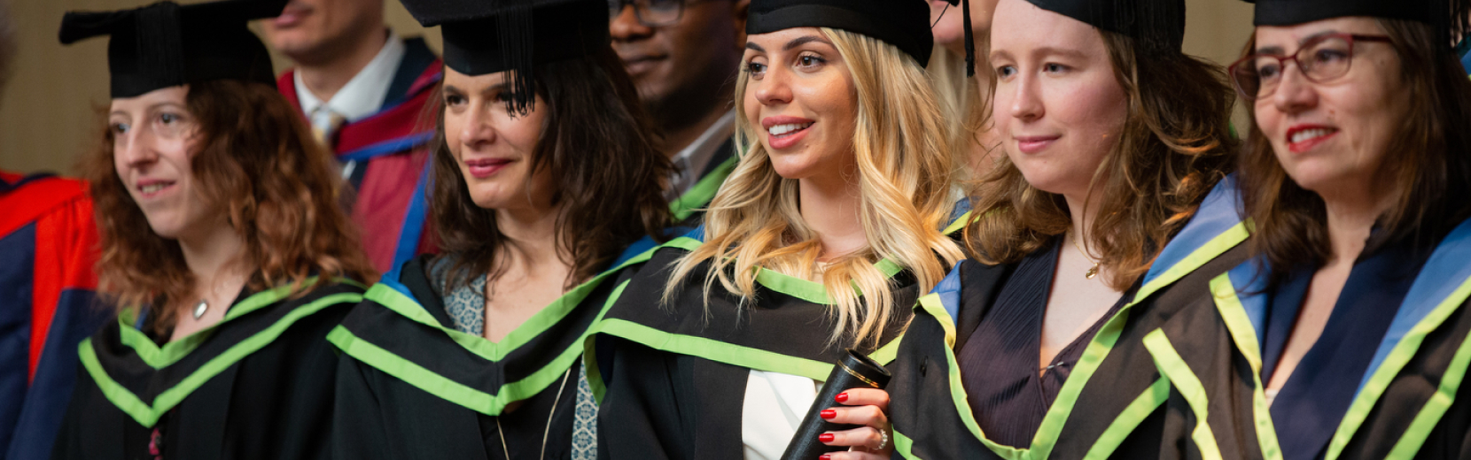 A group of graduates wearing caps and gowns stand in a line, smiling. Some are wearing glasses, and the attire features green and blue accents. The background is a soft blur, suggesting an indoor ceremony setting.