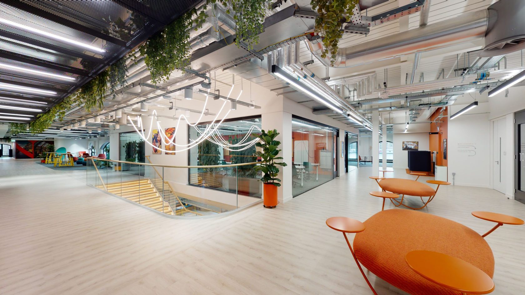 A modern office interior with open spaces, featuring a sleek design. Hanging plants and contemporary lighting add to the aesthetic. Orange seating areas and a staircase with glass railings are prominent, alongside exposed ducts and streamlined decor.
