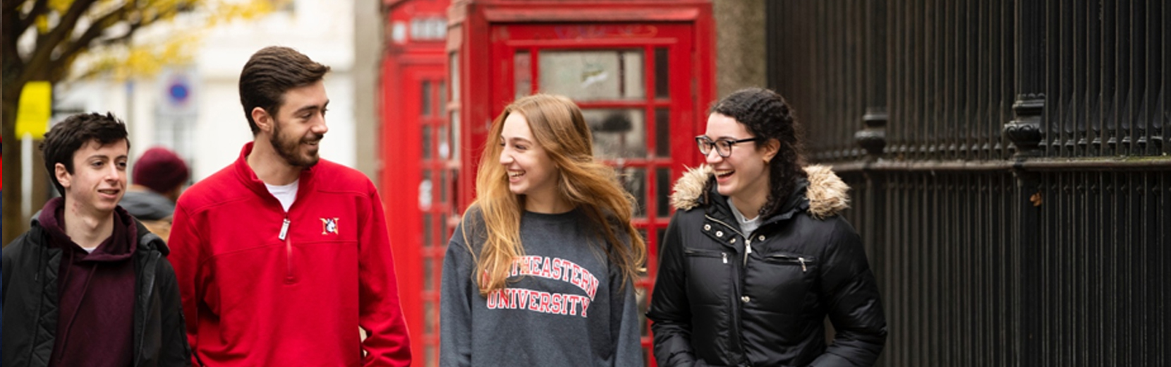 Four people are walking and talking, with two old-fashioned red phone booths in the background. They are wearing casual clothes and smiling, suggesting a relaxed atmosphere.