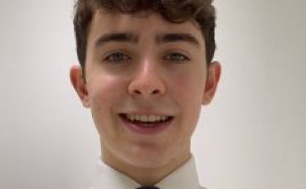 A young person with short, dark hair is smiling at the camera. They are wearing a white shirt and a dark tie. The background is plain and light-colored.