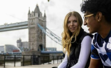 Two people sitting by the River Thames with the Tower Bridge in the background. One person, with long blonde hair, is smiling and wearing a dark vest. The other, with curly hair and glasses, is wearing a blue shirt with white patterns.