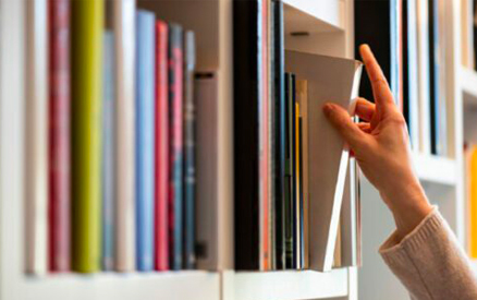 A hand reaches to pull a book from a bookshelf filled with colorful, upright books. The scene is indoors, with the focus on the action of selecting a book.