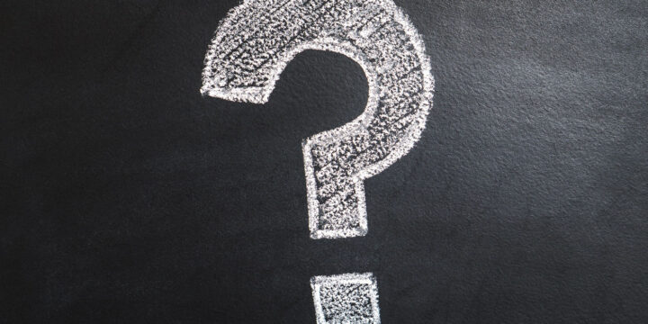 A large white chalk question mark is drawn on a blackboard, symbolizing inquiry or curiosity. The background is textured, emphasizing the chalk's slight smudging.