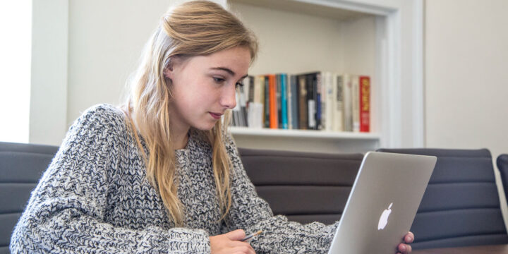 A person with long hair wearing a gray sweater is sitting at a table, focused on a laptop. There are books displayed on a shelf in the background.
