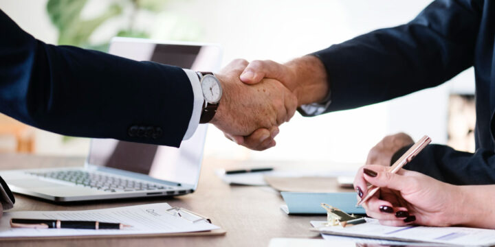 Two people in business attire are shaking hands over a desk with documents, a laptop, and writing materials. One person is taking notes with a pen, suggesting a professional meeting or agreement.