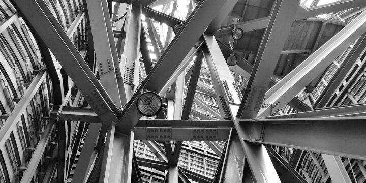 A black and white image showing the intricate steel framework of a modern architectural structure. Numerous beams and joints intersect at various angles, creating a complex, geometric pattern.