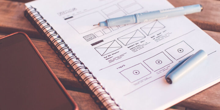 A spiral notebook open to a page with hand-drawn website wireframes and designs rests on a wooden table. A black smartphone and two light blue pens are nearby, suggesting a workspace for planning and sketching digital layouts.