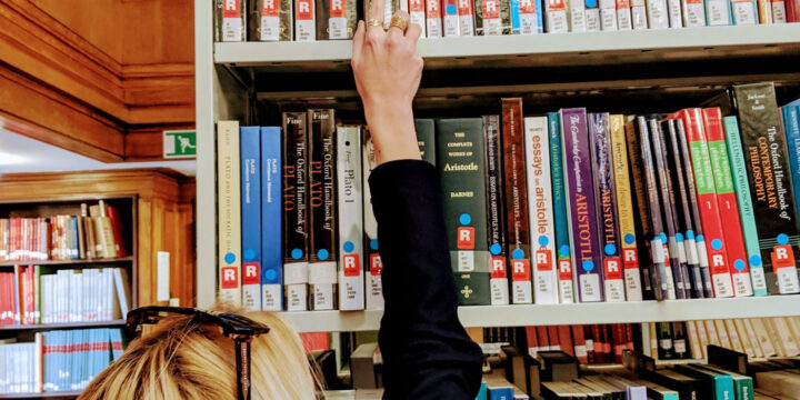 A person reaches for a book on a high library shelf filled with various books. The person has blonde hair and wears sunglasses on their head. The library has wooden accents and several books are marked with red and blue labels.