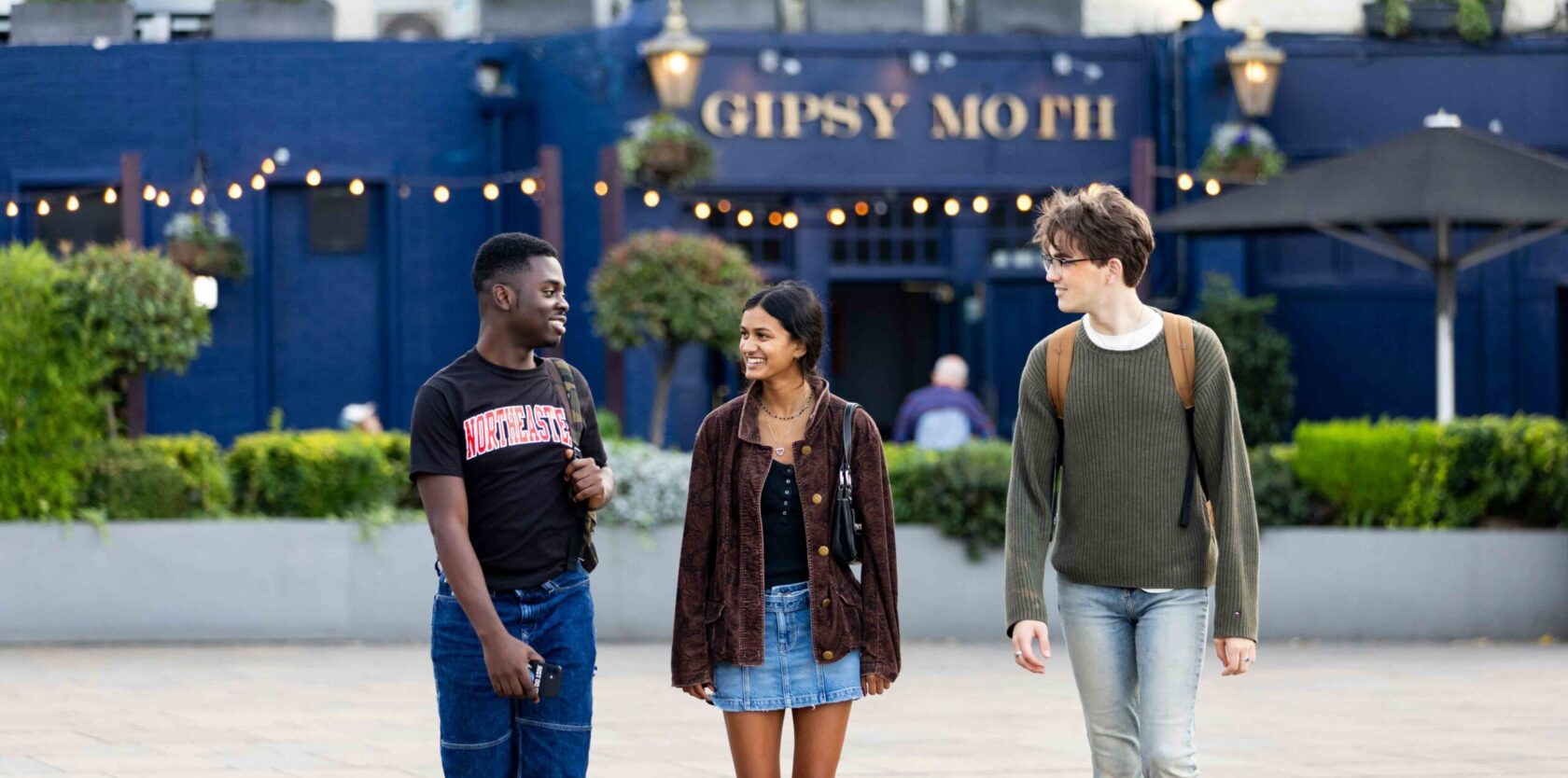Three young adults walk together outside a building labeled "Gipsy Moth." They are smiling and appear to be having a conversation. The building has string lights and greenery in the foreground.