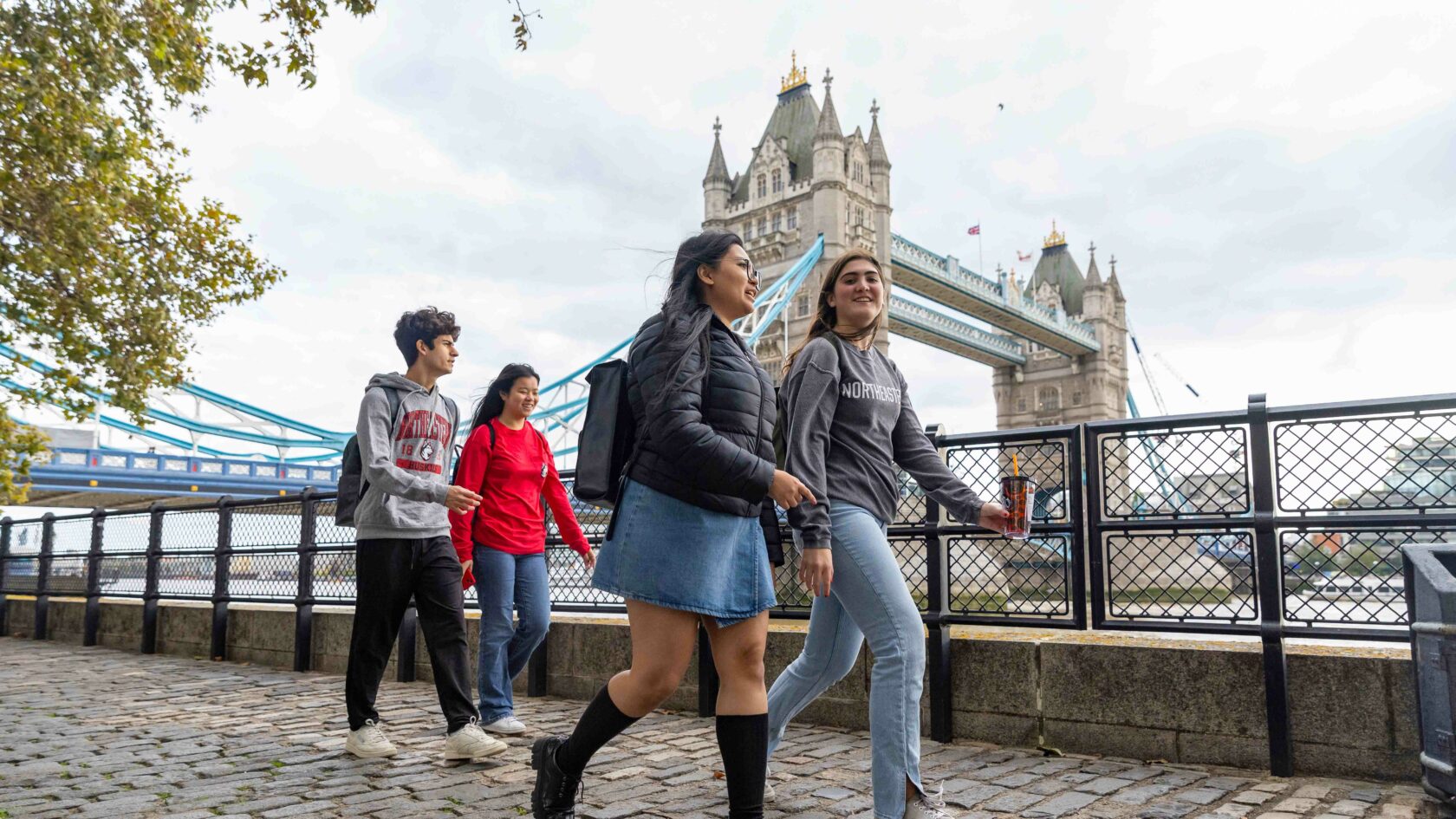 A group of four people walks along a cobblestone path by the river, with the Tower Bridge in the background. They appear to be talking and enjoying the day, under a cloudy sky.