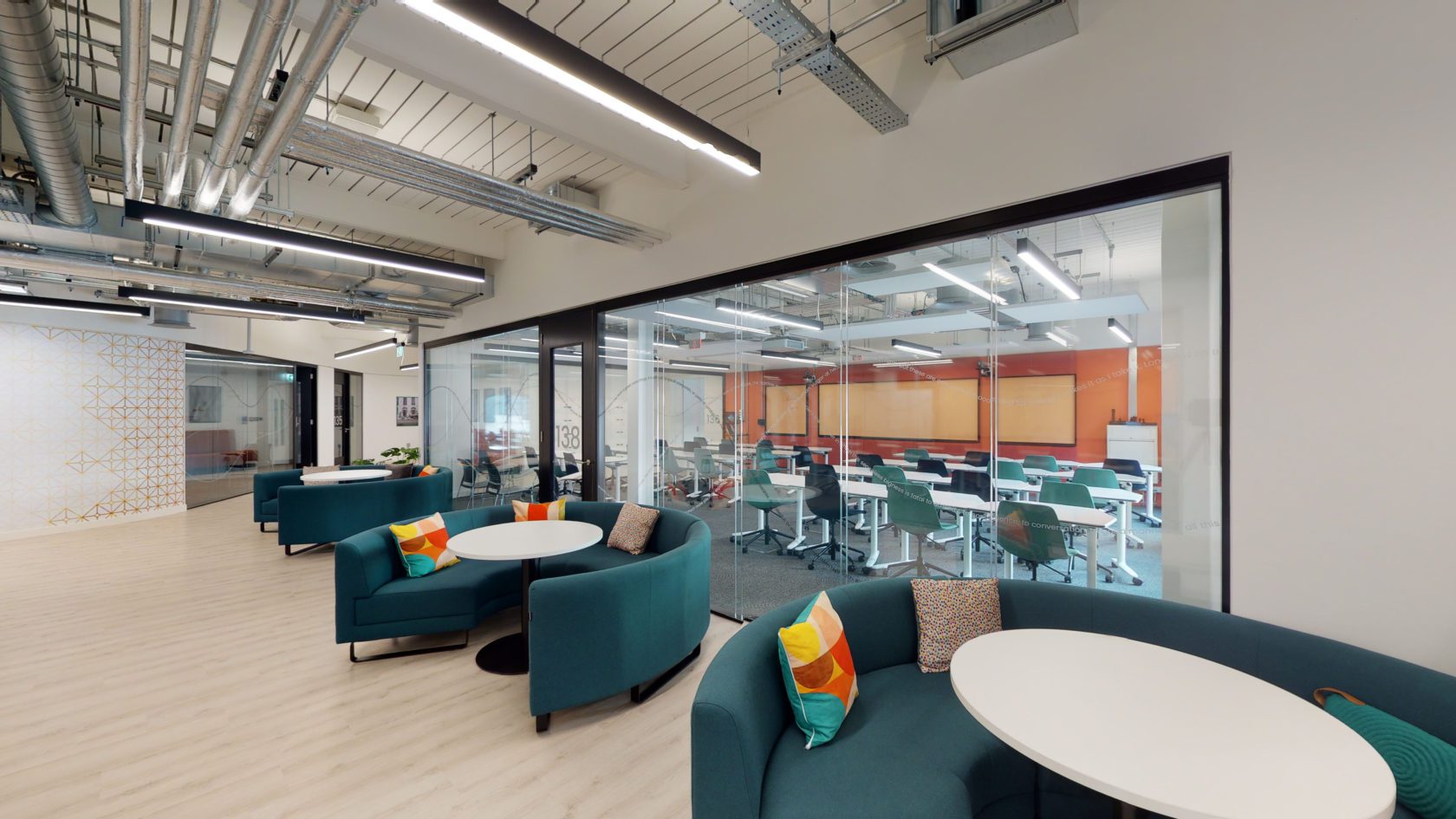 Modern office space with teal armchairs and round white tables in the foreground. Glass wall separates a conference room with desks and chairs, vibrant orange accent wall, and visible ceiling pipes. Bright lighting and wooden flooring throughout.
