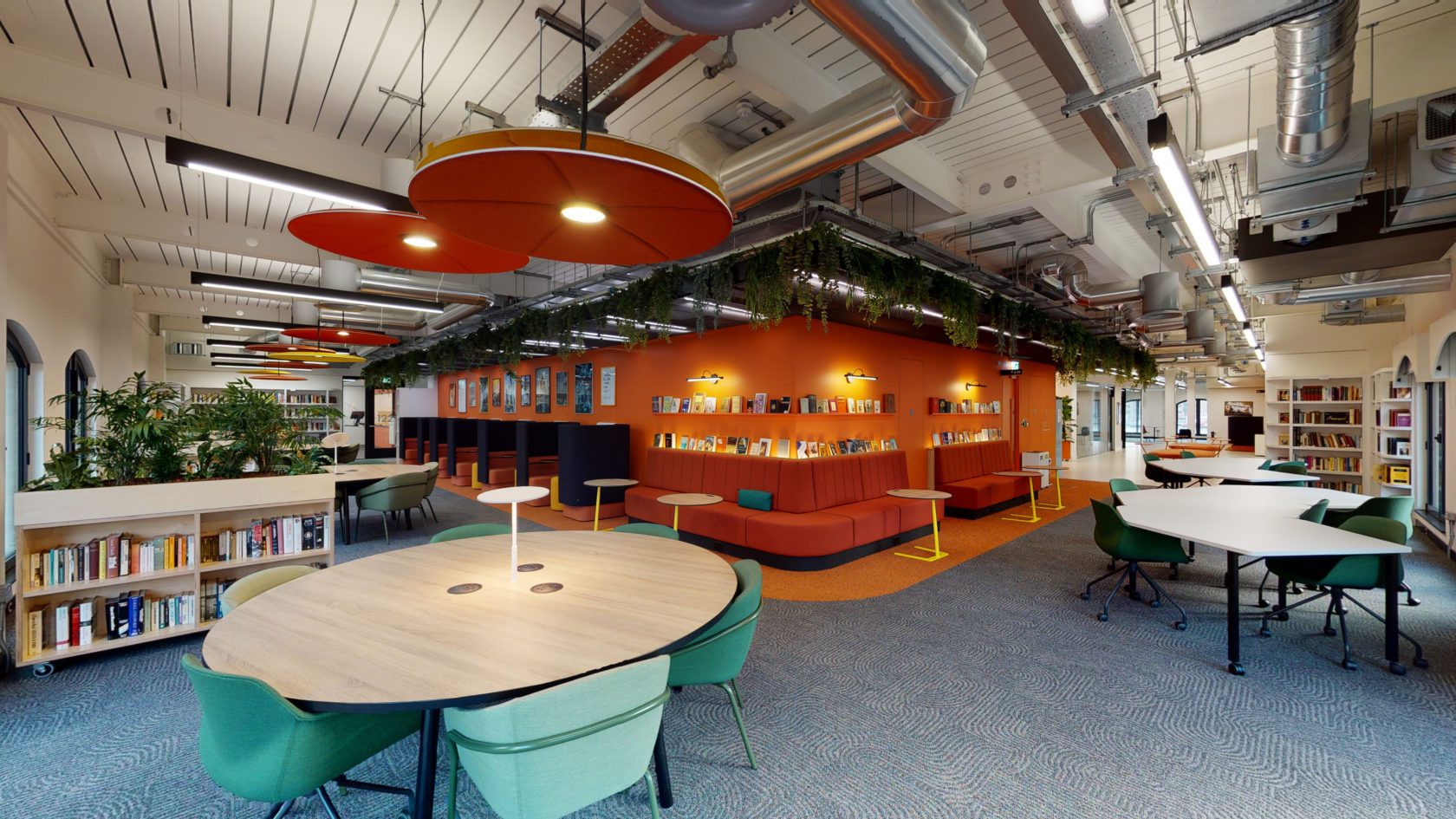 A modern, brightly lit library interior with circular tables and green chairs in the foreground. Orange walls feature bookshelves and seating. Exposed ducts and plants hang from the ceiling, creating a vibrant and open space.