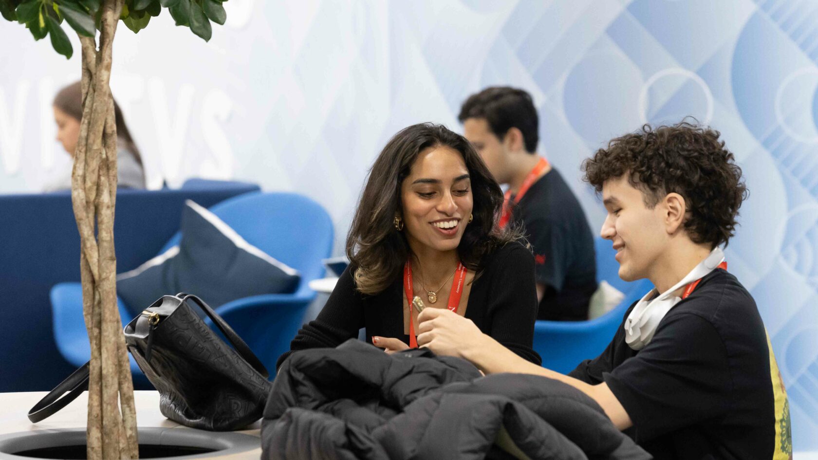 Two people sitting at a table, smiling and engaged in conversation. The setting appears casual, with a potted plant near them and patterned blue walls in the background. They are wearing lanyards and there is a backpack and jacket on the table.
