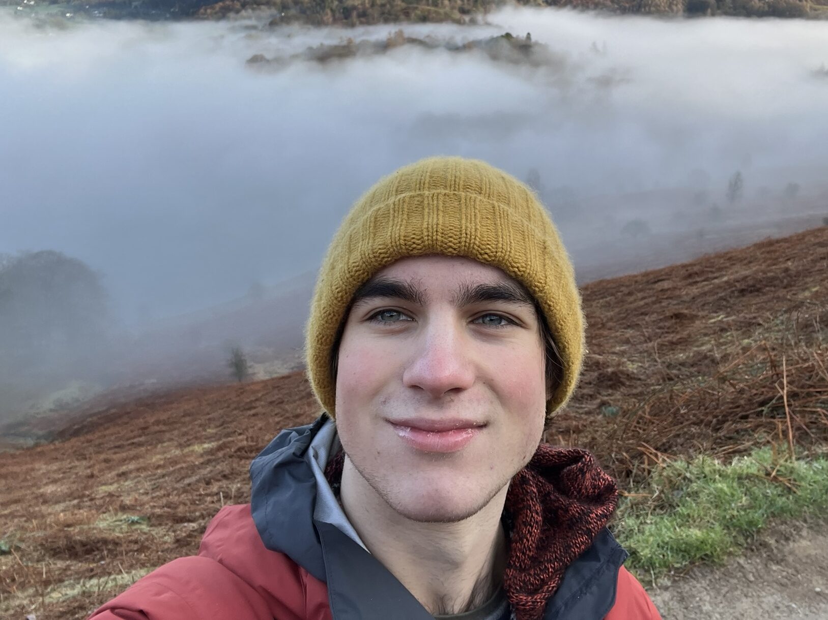 A person wearing a mustard yellow beanie and red jacket takes a selfie on a foggy hillside. The background features mist-covered hills and sparse vegetation.