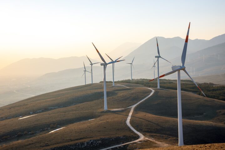 Wind turbines stand on rolling hills under a clear sky at sunset, blending culture and technology amid mountains in the background and a winding dirt road leading through the landscape.