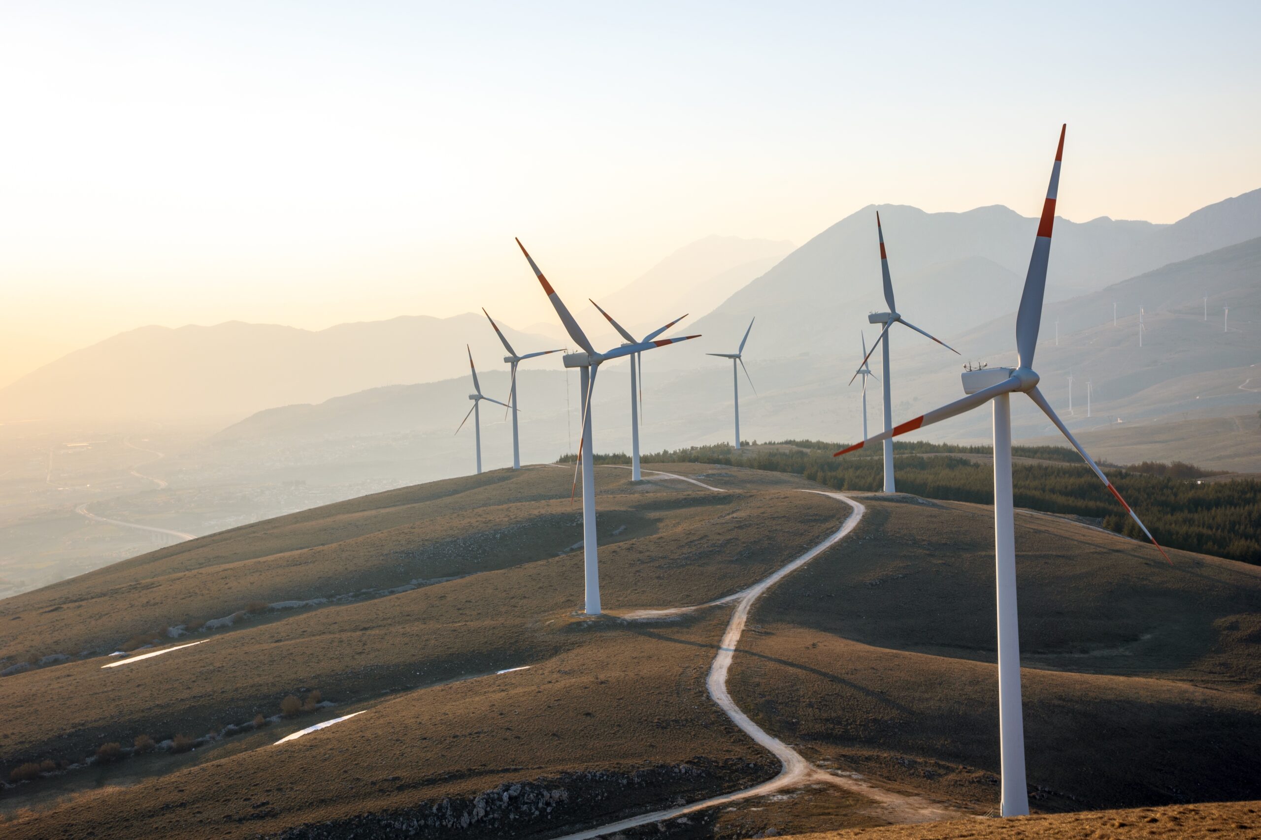 Wind turbines stand on rolling hills under a clear sky at sunset, blending culture and technology amid mountains in the background and a winding dirt road leading through the landscape.