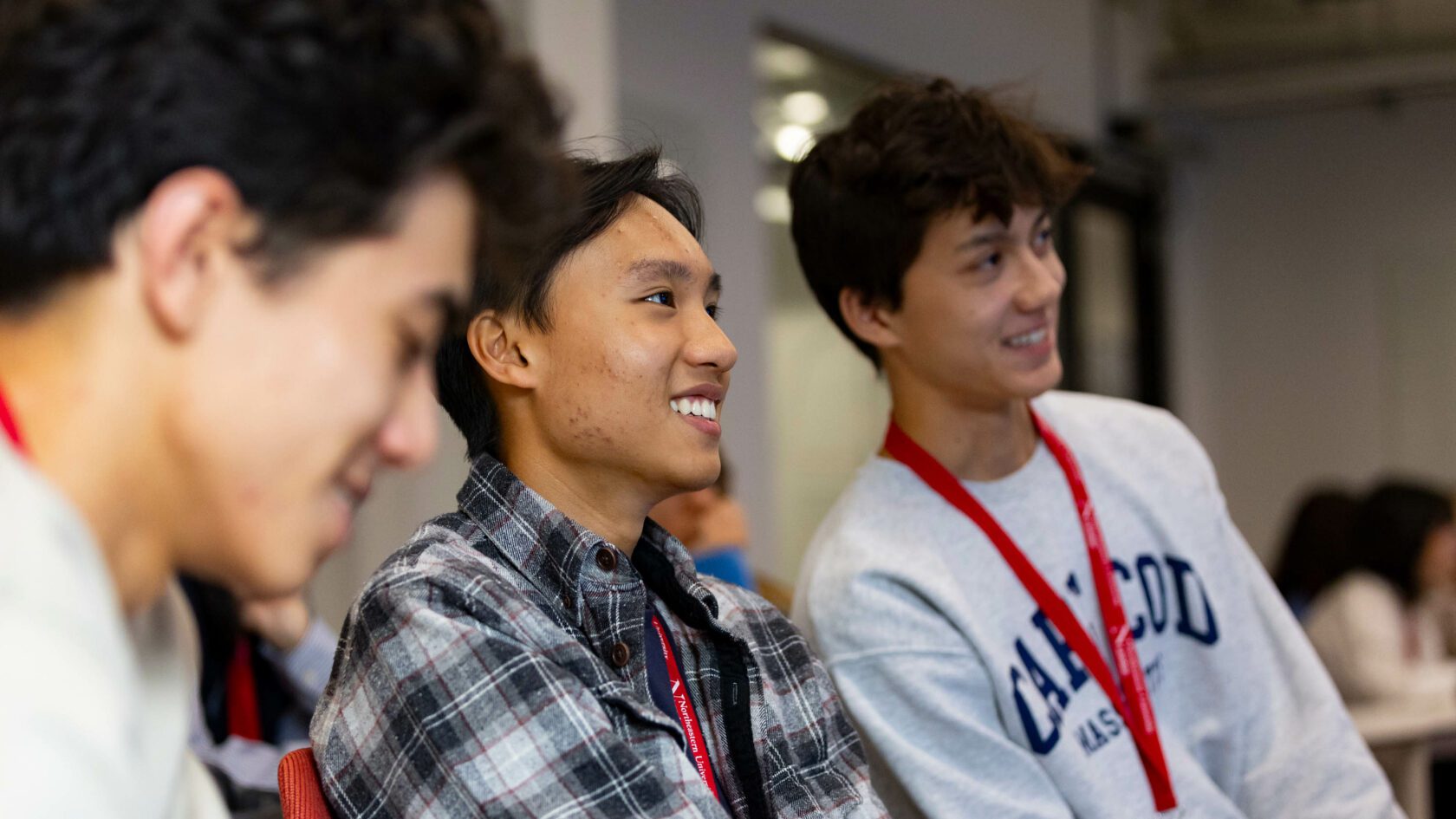 Three young men sit together in a classroom, wearing casual clothing with red lanyards. They appear engaged and smiling, suggesting they are listening to a presentation or discussion.