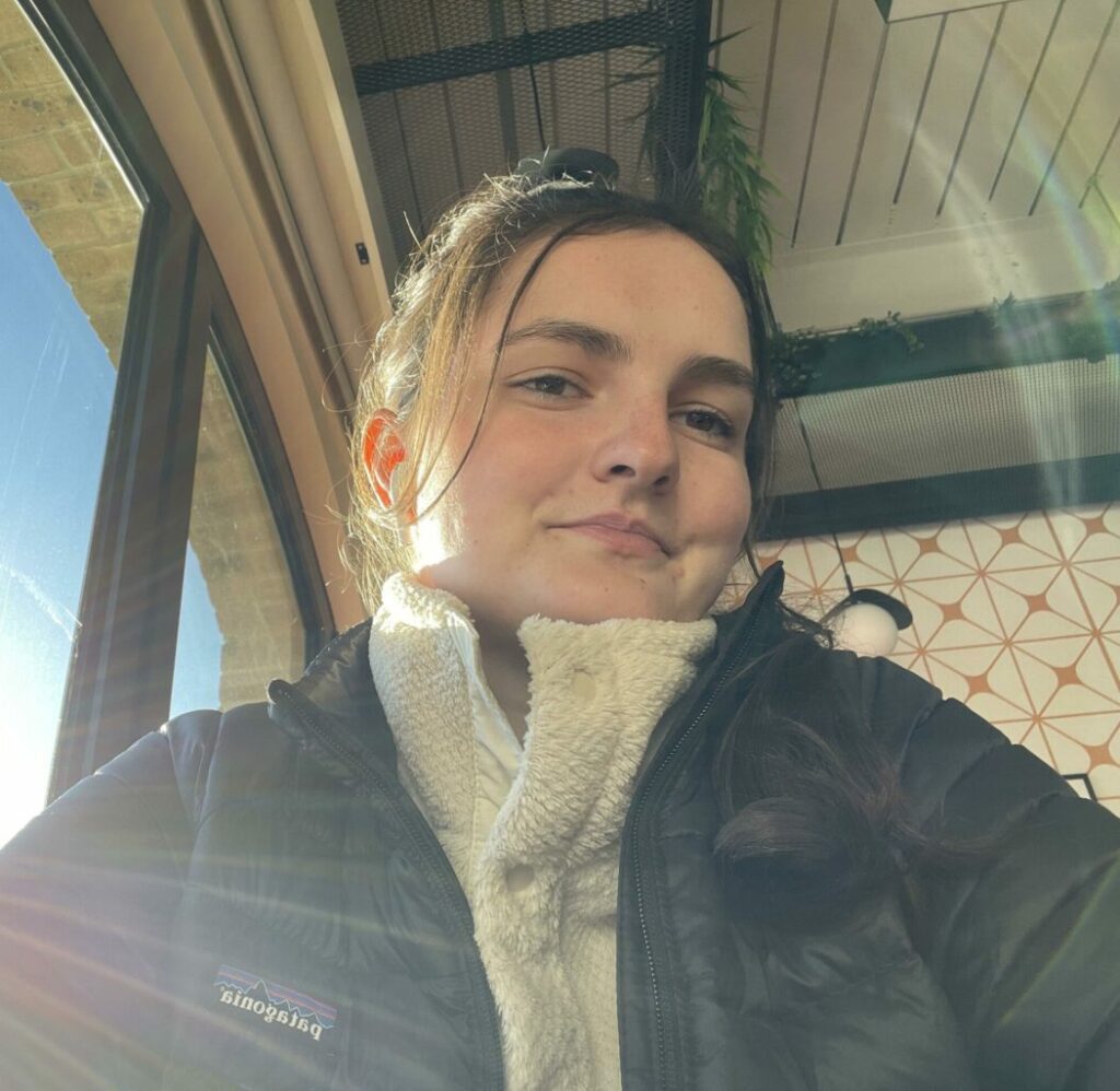 A person in a warm jacket and scarf sits indoors, smiling slightly. Sunlight streams in from the window, creating a cozy atmosphere. The ceiling has some hanging plants and geometric patterns on the walls add a stylish touch.