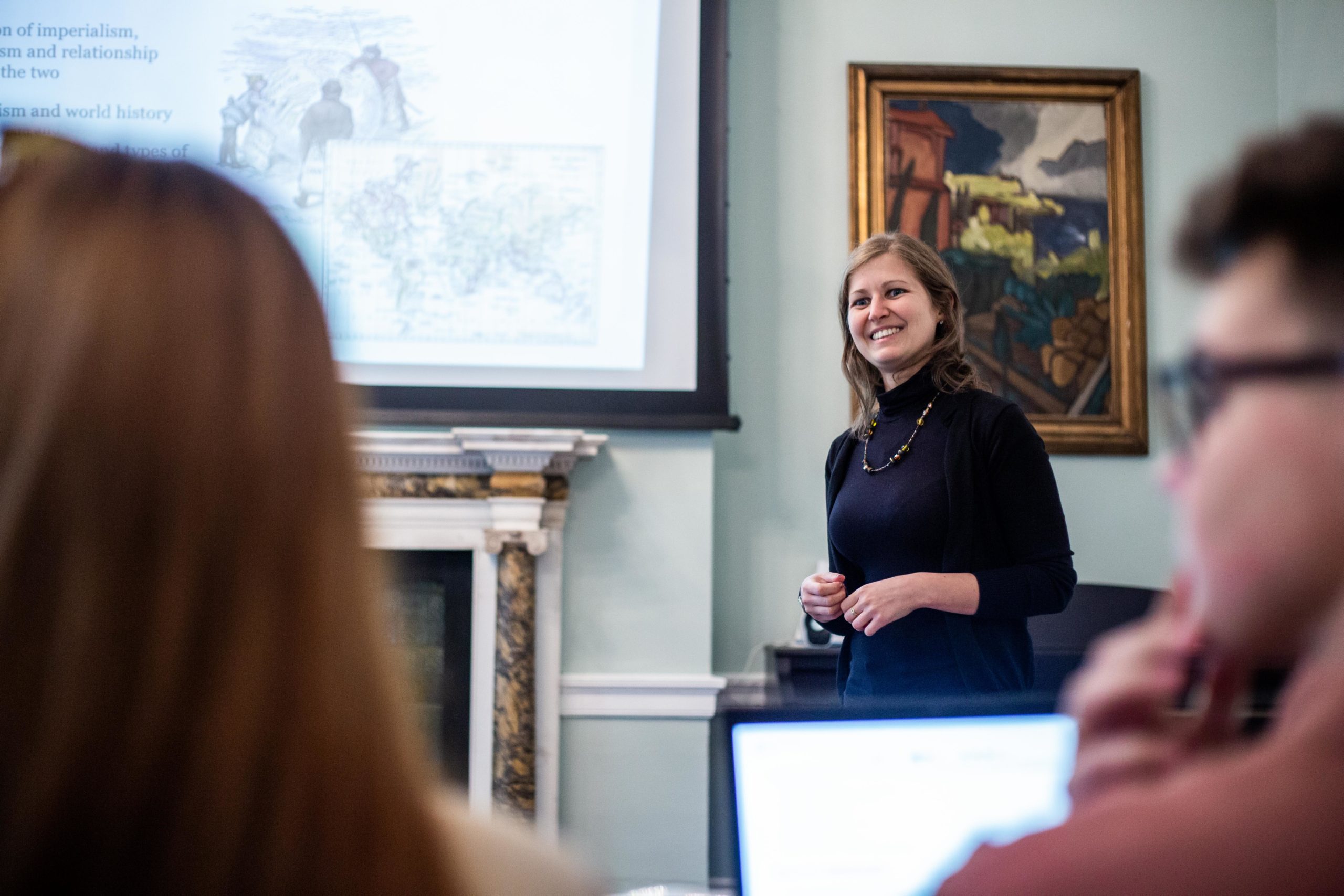 An undergraduate woman stands smiling in front of a classroom, presenting with a projector screen displaying historical maps and images. Students sit facing her, engaged. The room exudes traditional decor with framed art on the walls.