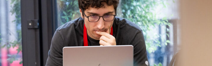 A person with curly hair and glasses is intently looking at a laptop screen. They are sitting indoors, near a window with a view of greenery outside. They rest their hand on their chin, appearing deep in thought.
