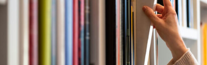 A hand reaches for a book on a colorful bookshelf, with vertical spines in varying shades of pink, blue, green, and more. The person appears to be selecting or organizing the books.