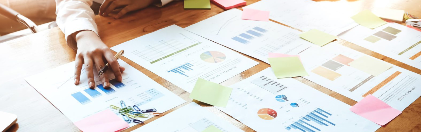 A person's hand organizes various financial documents and colorful sticky notes spread across a wooden table. Some documents display graphs and charts, and a few paperclips are visible on the table.