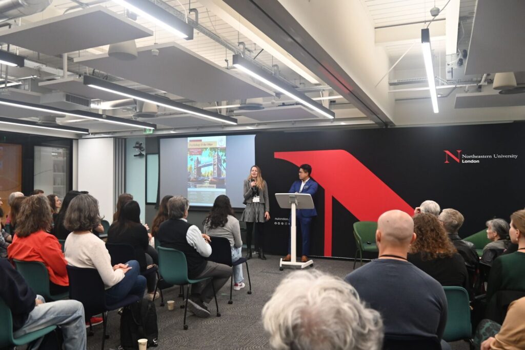 A woman stands at a podium speaking to a seated audience in a modern conference room. A presentation is displayed on a screen behind her. Another person stands nearby. The room features industrial-style lighting and a logo on the wall.