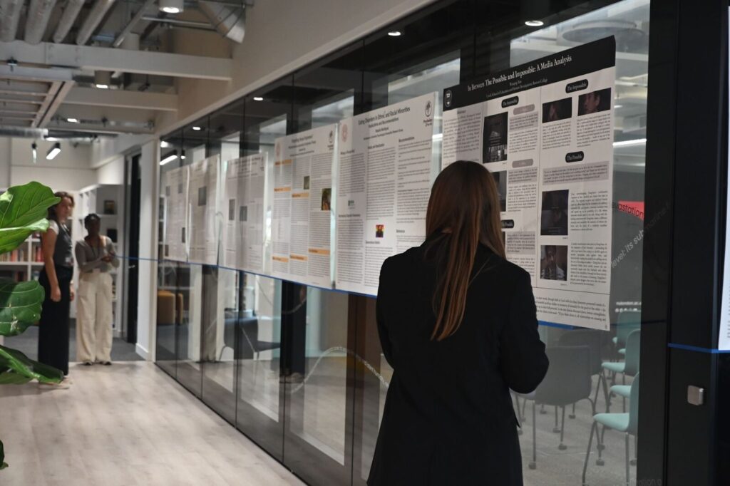 A woman stands in a hallway, closely examining a series of scientific posters displayed on a glass wall. The setting appears to be an academic or professional conference, with other attendees visible in the background.