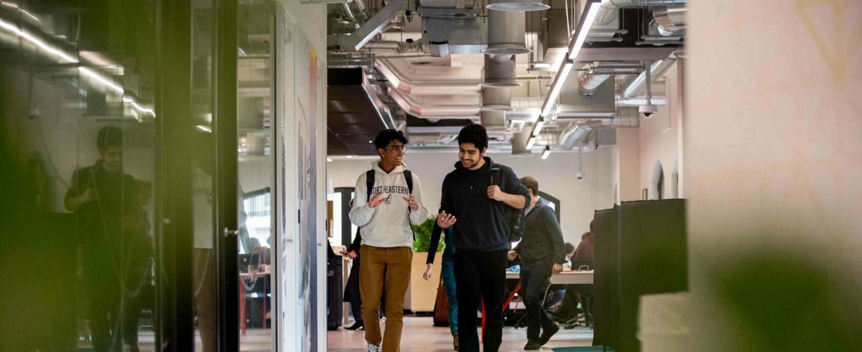 Two people walk and chat in a modern office hallway with visible ductwork and lighting overhead. Others can be seen working in the background. The scene is framed by blurred foliage in the foreground.