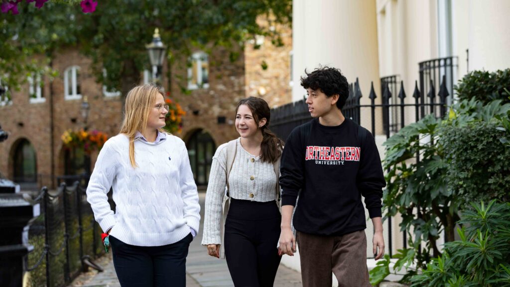 Three people walking down a sidewalk beside a building with ornate fencing and greenery. One wears a white sweater, another a beige cardigan, and the third a black sweatshirt with "Northeastern University" written on it. Trees and a lamppost are in the background.