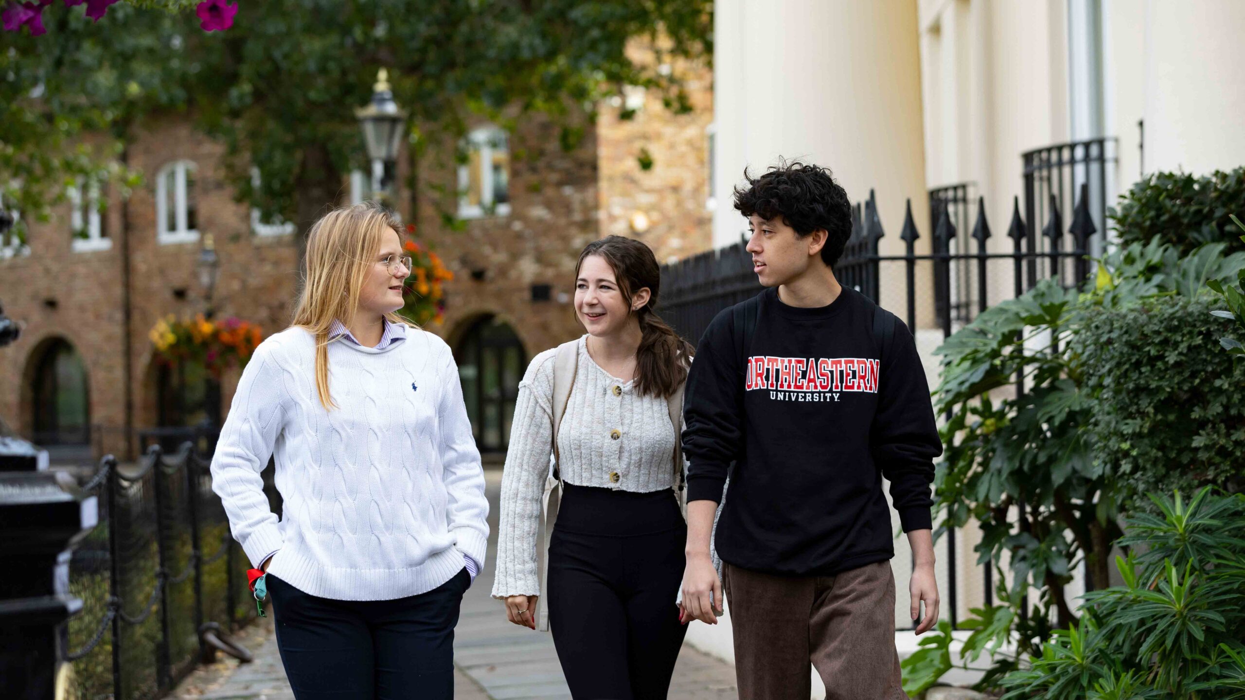 Three people walking down a sidewalk beside a building with ornate fencing and greenery. One wears a white sweater, another a beige cardigan, and the third a black sweatshirt with "Northeastern University" written on it. Trees and a lamppost are in the background.