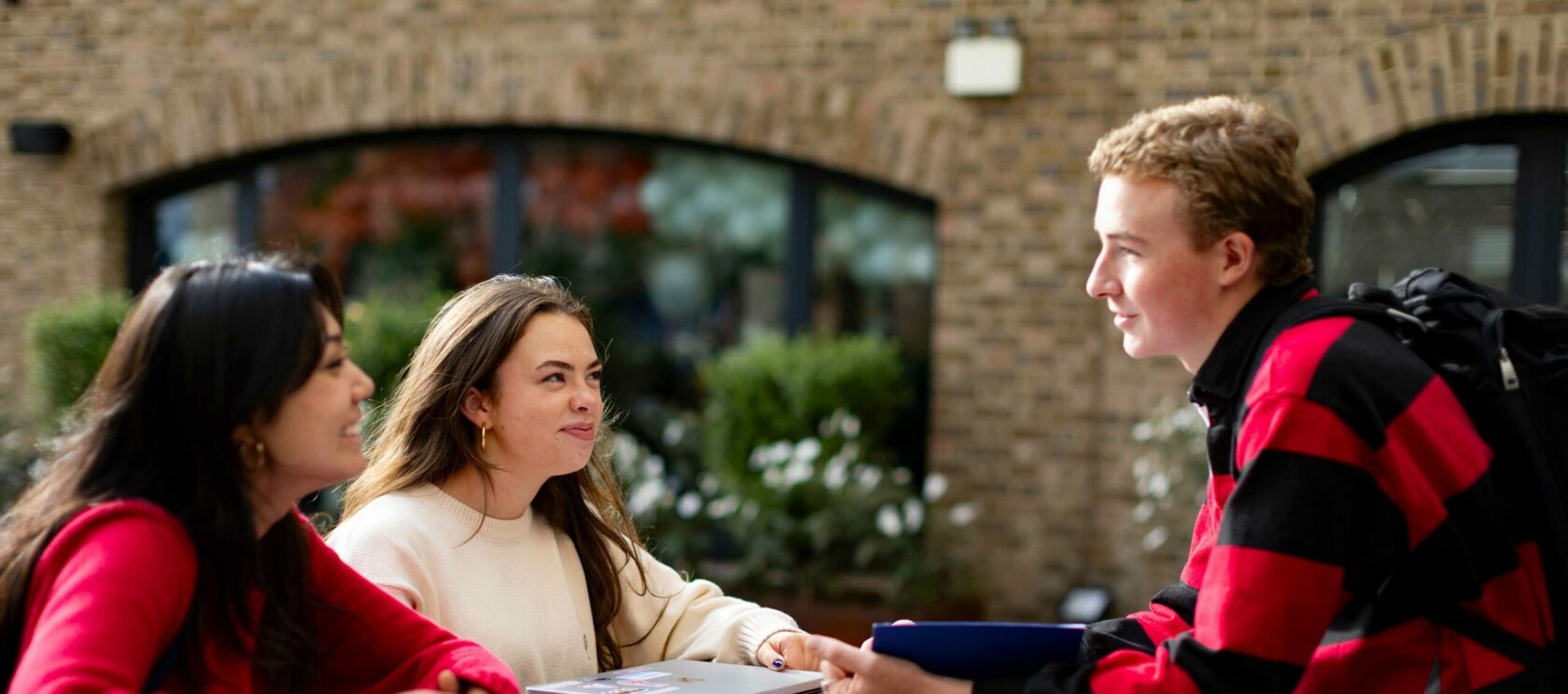 Three young adults sit at an outdoor table engaged in conversation. Two are on one side, facing a young man in a red and black striped shirt. A brick building and some greenery are in the background.