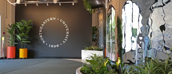 A modern hallway with plants and mirrors, featuring a black wall with the Northeastern University logo and the year 1898 in white text.