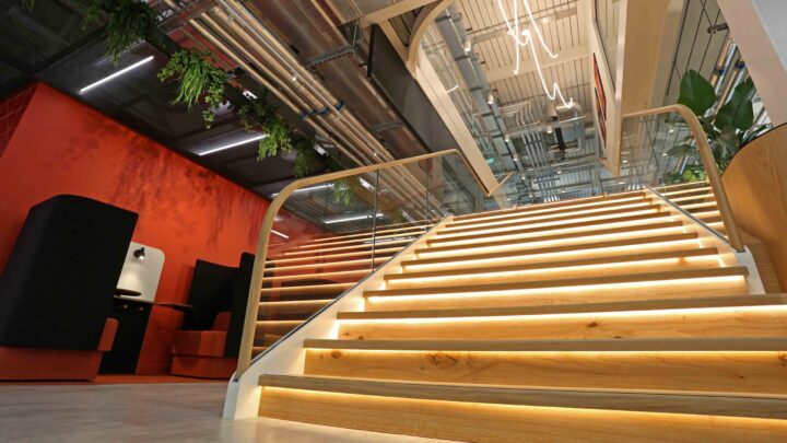 Wide, modern indoor staircase with wooden steps and yellow lighting underneath each step, leading up to an industrial-style ceiling with exposed pipes and hanging plants. Contemporary furniture is visible to the left.