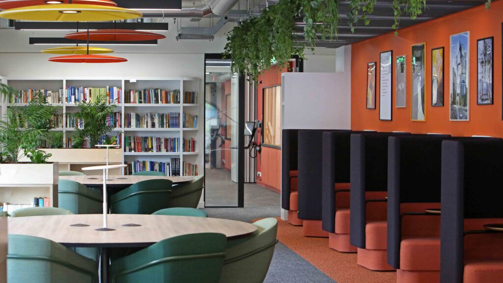 Modern office space with cozy seating booths, green chairs at round tables, bookshelves, orange accent wall with framed pictures, and hanging plants from the ceiling.
